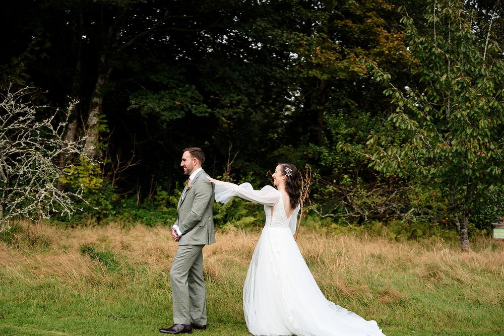 A couple in wedding attire playfully dance and twirl in a scenic outdoor field with trees in the background.