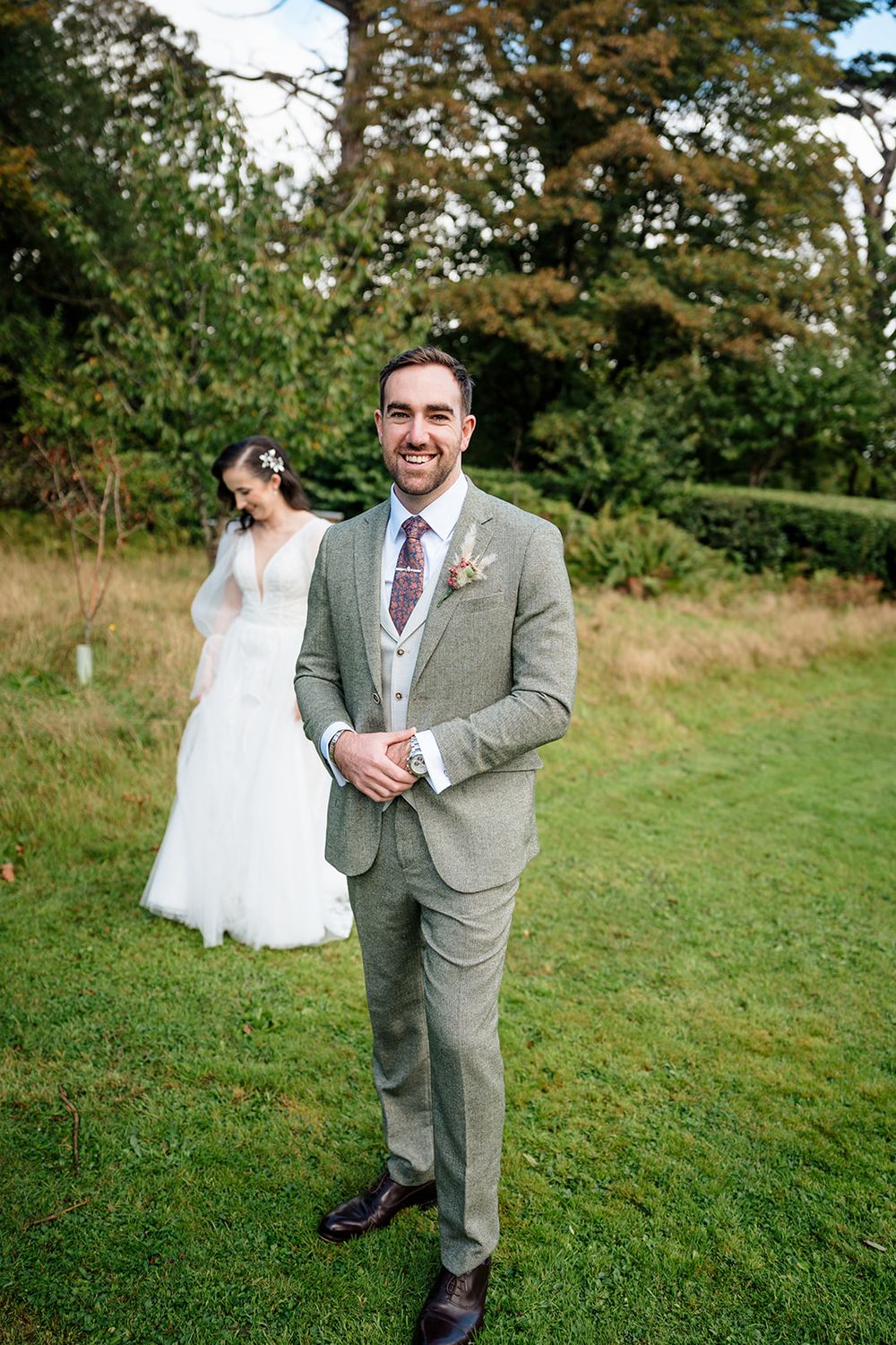 Wedding photo showing a couple outdoors with the groom in a gray suit and the bride in white against a natural backdrop.