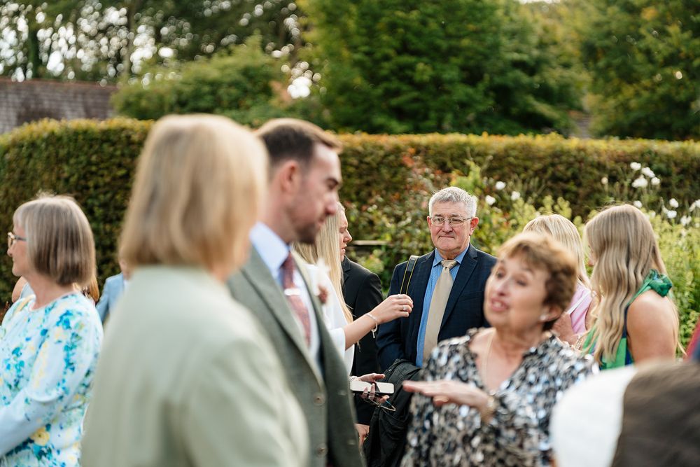 Outdoor garden party scene with guests mingling among green hedges and trees.