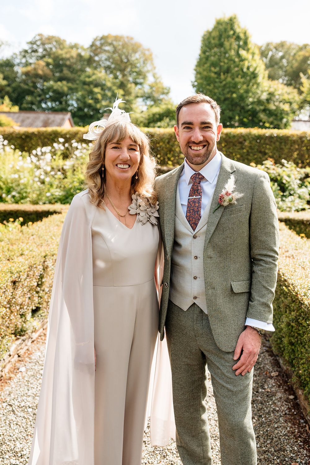 A happy couple in wedding attire laughs together in a sunny garden setting wearing beige and sage green formal wear.