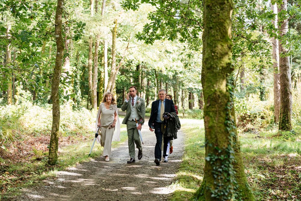 Wedding party walks along a tree-lined path in a lush green forest setting during a summer celebration.