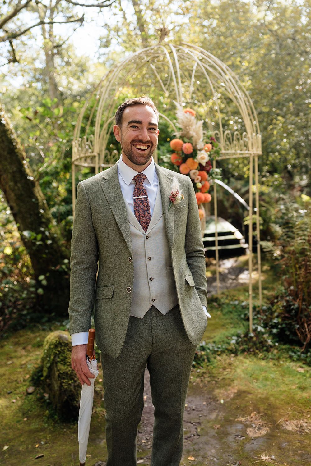 A groom in a stylish grey tweed suit and vest stands smiling in front of a floral wedding arch in a garden setting.