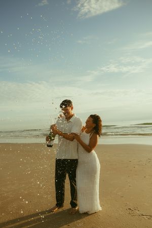 Couple celebrating on beach, popping champagne bottle, woman in white dress, ocean backdrop, golden hour light.