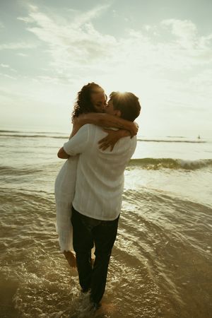 Couple embracing and kissing on a sunny beach with waves, backlit by golden sunlight, wearing white and black.