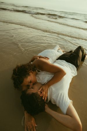 Romantic couple lying on a sandy beach shore, embracing and kissing as gentle ocean waves wash around them.