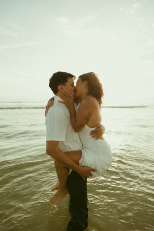 Couple sharing a romantic kiss while standing in ocean waves, man lifting woman in white dress at sunset.