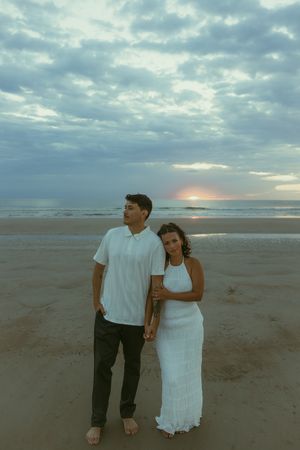 Couple in white outfits holding hands on a sandy beach at sunset with dramatic cloudy sky backdrop.