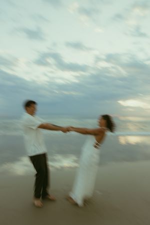 Couple dancing romantically on a beach at sunset, holding hands in motion blur with ocean waves in the background.