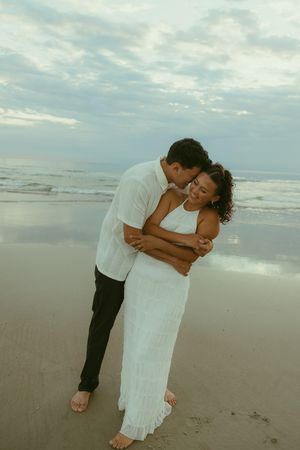 Couple embracing on a beach at dusk, woman in white dress, man in white shirt, with ocean waves in the background.