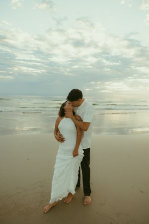 Couple sharing a romantic kiss on a sandy beach at sunset, woman in white dress, man in white shirt and dark pants.