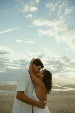 Romantic couple embracing and almost kissing on a sandy beach at sunset with a cloudy blue sky backdrop.