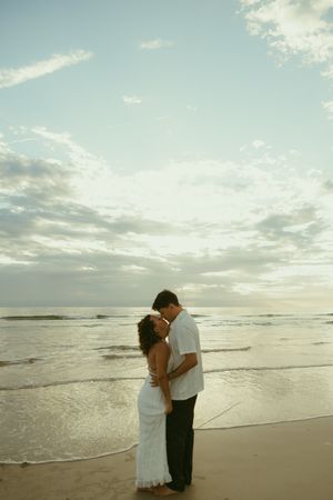 Romantic couple embracing on a beach at sunset, woman in white dress, waves and cloudy sky in background.