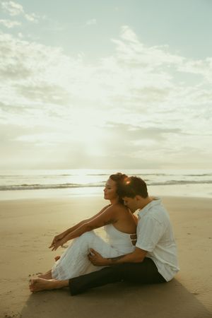 A couple sits together on a sandy beach at sunset, the woman in a white dress leaning back against her partner.