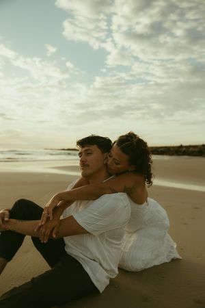 A couple sits on a sandy beach at sunset, the woman in white embracing the man from behind near the ocean waves.