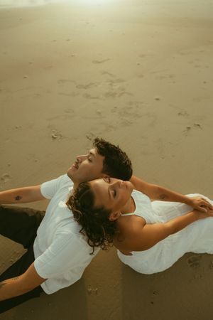 Couple lying back-to-back on sandy beach, wearing white, gazing upward in warm golden light.