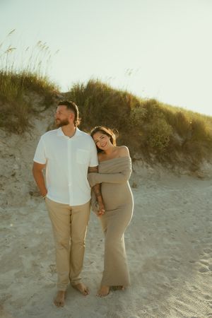 Couple embracing on sandy beach dunes at golden hour, woman in tan dress leaning on man in white shirt.