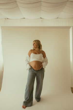 Pregnant woman in white crop top and gray wide-leg jeans cradling baby bump in a bright studio setting.