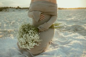 Pregnant woman in beige knit dress sitting on beach sand holding white wildflower bouquet at golden sunset.