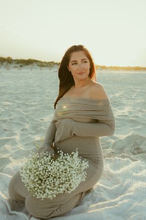 Pregnant woman in beige off-shoulder knit dress sitting on sandy beach holding white baby's breath flowers at golden sunset.