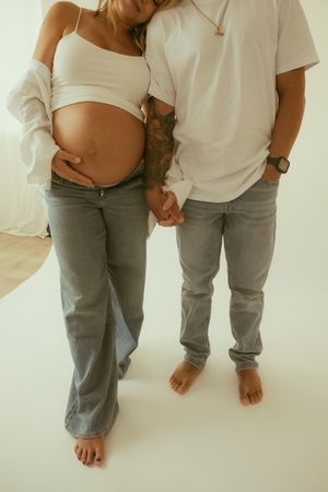 Pregnant couple holding hands, woman in white crop top and jeans cradling baby bump, man with tattoos in white tee.