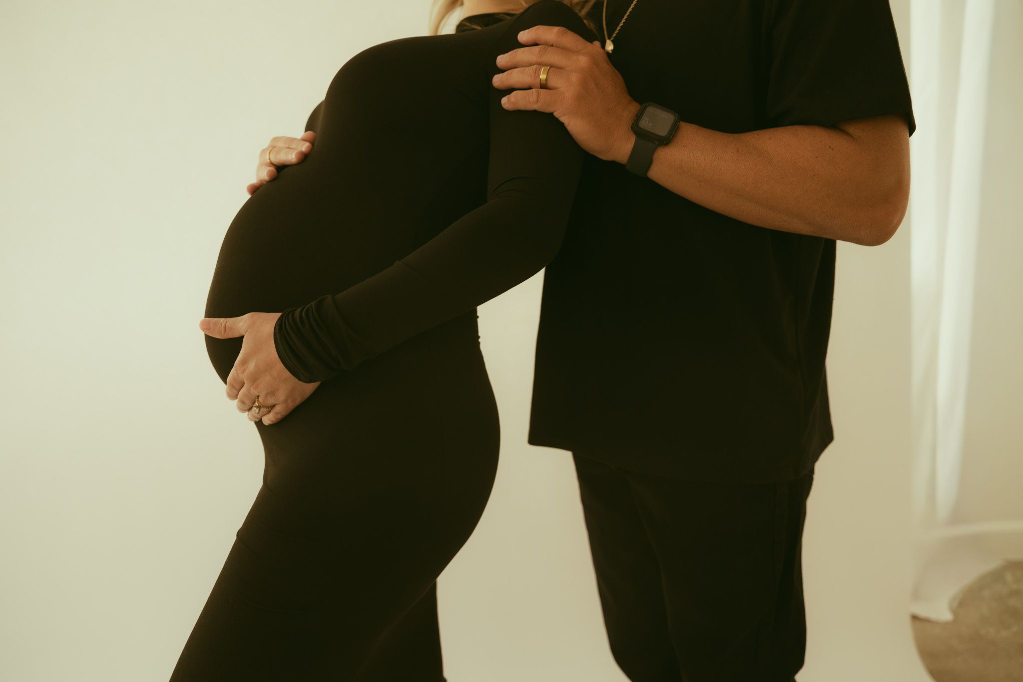 Pregnant woman in black dress cradling baby bump with partner standing behind her in a bright studio.