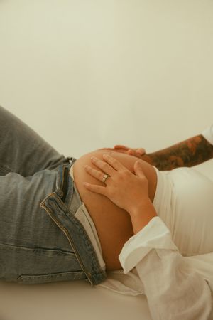Close-up of pregnant woman's baby bump with ring on finger, wearing white shirt and jeans.
