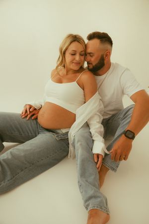 Couple sitting on white floor, pregnant woman in white crop top and jeans, man in white shirt embracing her tenderly.