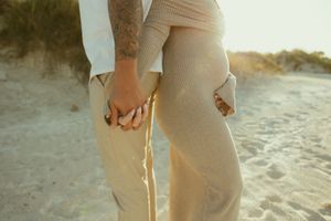 Couple holding hands on a sandy beach at golden hour, one partner showing a tattooed arm.