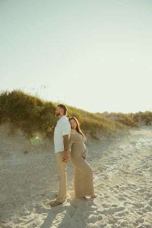 Pregnant couple holding hands on sandy beach dunes at golden hour, woman in tan maxi dress leaning against partner.