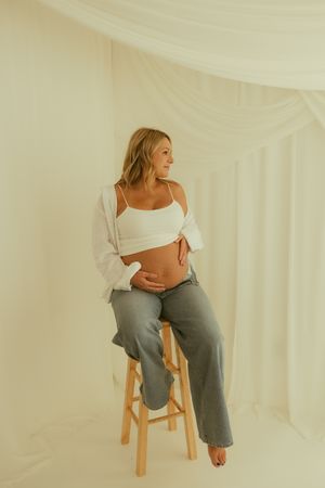 Pregnant woman in white crop top and gray pants sits on wooden stool, cradling baby bump in bright studio.