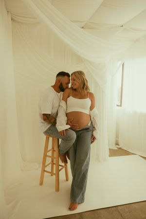 A couple poses tenderly in a white draped studio, the pregnant woman cradling her baby bump in a white crop top and jeans.