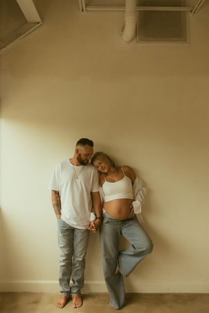 A couple poses against a beige wall, the pregnant woman in a white crop top leaning on her partner who wears a white tee and jeans.