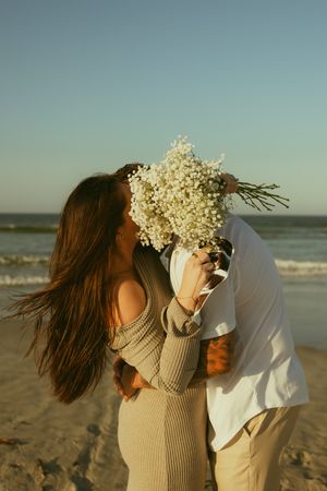 Couple kissing behind white wildflower bouquet on sandy beach at sunset, woman in knit dress with flowing hair.