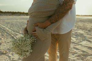 Pregnant couple embracing on a sandy beach at sunset, woman holding white baby's breath flowers, man with tattooed arms.
