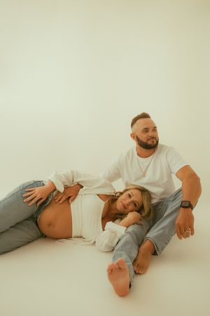 Couple in white tops and jeans posing on white floor, pregnant woman lying with head on partner's lap.