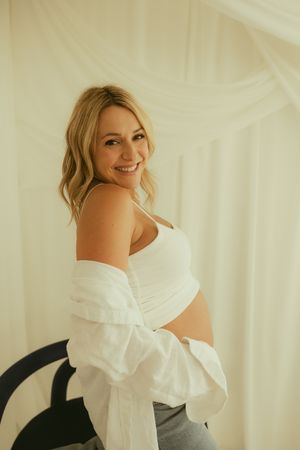 Smiling pregnant woman in white crop top and open shirt showing baby bump, maternity photo session.