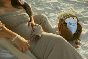Pregnant woman in beige ribbed dress relaxing on sandy beach with a floral bouquet beside her.