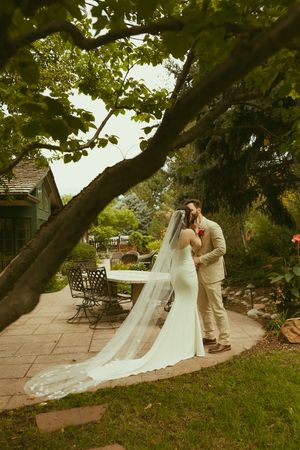 Bride and groom share a romantic moment in a lush garden, her cathedral veil trailing on a stone patio.