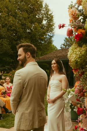 Bride in white strapless gown and groom in tan suit stand at outdoor wedding altar adorned with colorful floral arrangements.