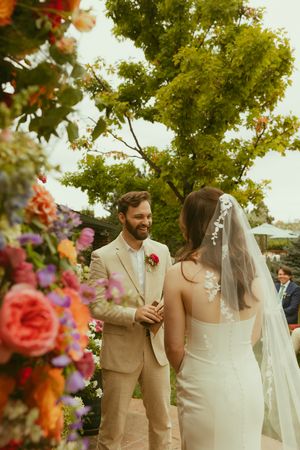 Groom in tan suit smiles at bride in lace wedding dress and veil during outdoor ceremony with colorful floral arrangements.