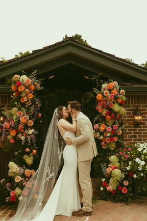 Bride and groom share first kiss under vibrant floral arch with colorful blooms at outdoor wedding ceremony.