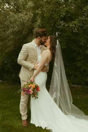 A bride and groom share a romantic kiss outdoors, she holds a colorful bouquet and wears a flowing veil.