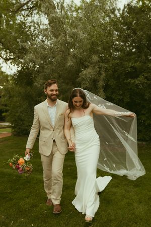 Bride in white gown with flowing veil walks hand-in-hand with groom in tan suit holding colorful bouquet outdoors.