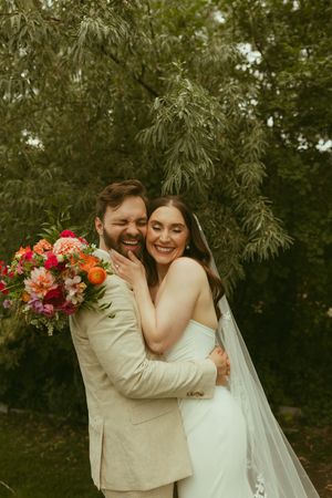 A joyful bride and groom embrace outdoors, she holds a vibrant pink and orange bouquet, wearing a white gown with veil.