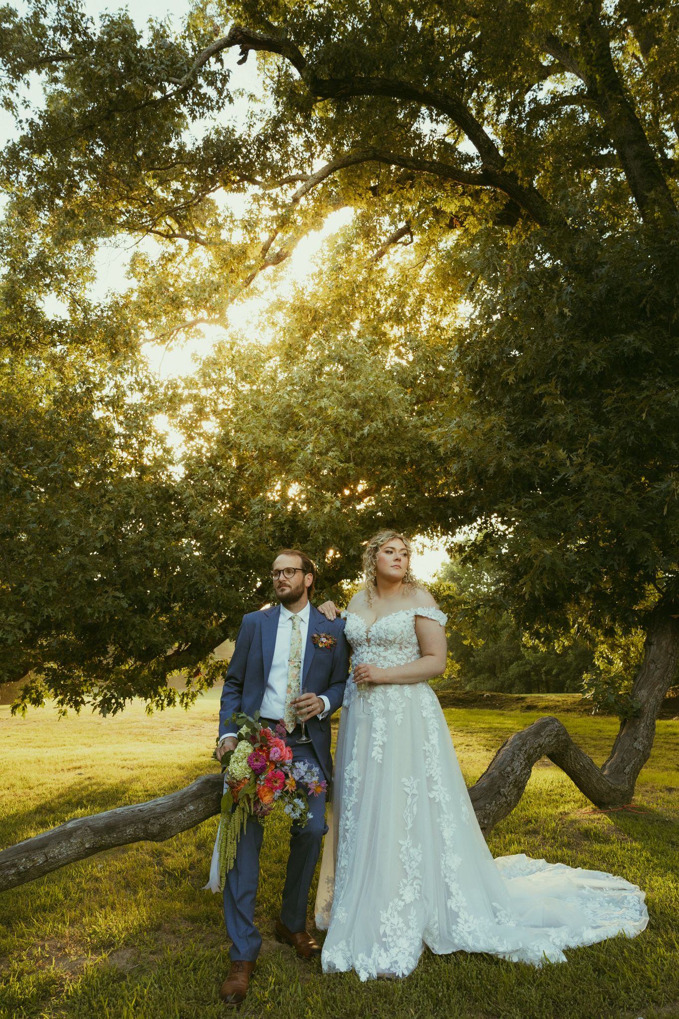 Bride and groom pose under a sunlit oak tree in a lush green field, bride in lace gown, groom in blue suit.