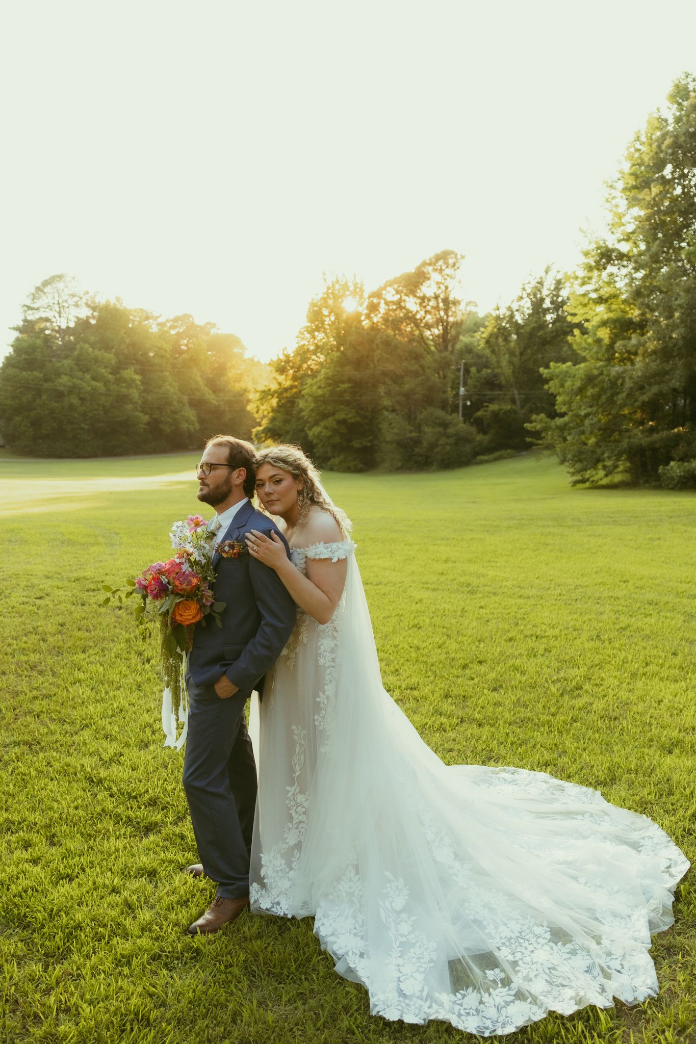 Bride in lace gown leans on groom in gray suit holding colorful bouquet at golden hour on green lawn.