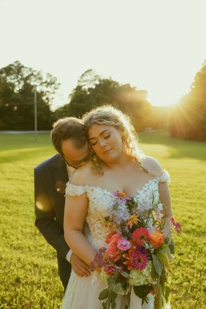 Bride and groom share a tender golden hour moment outdoors, bride holding a vibrant bouquet of pink and white flowers.