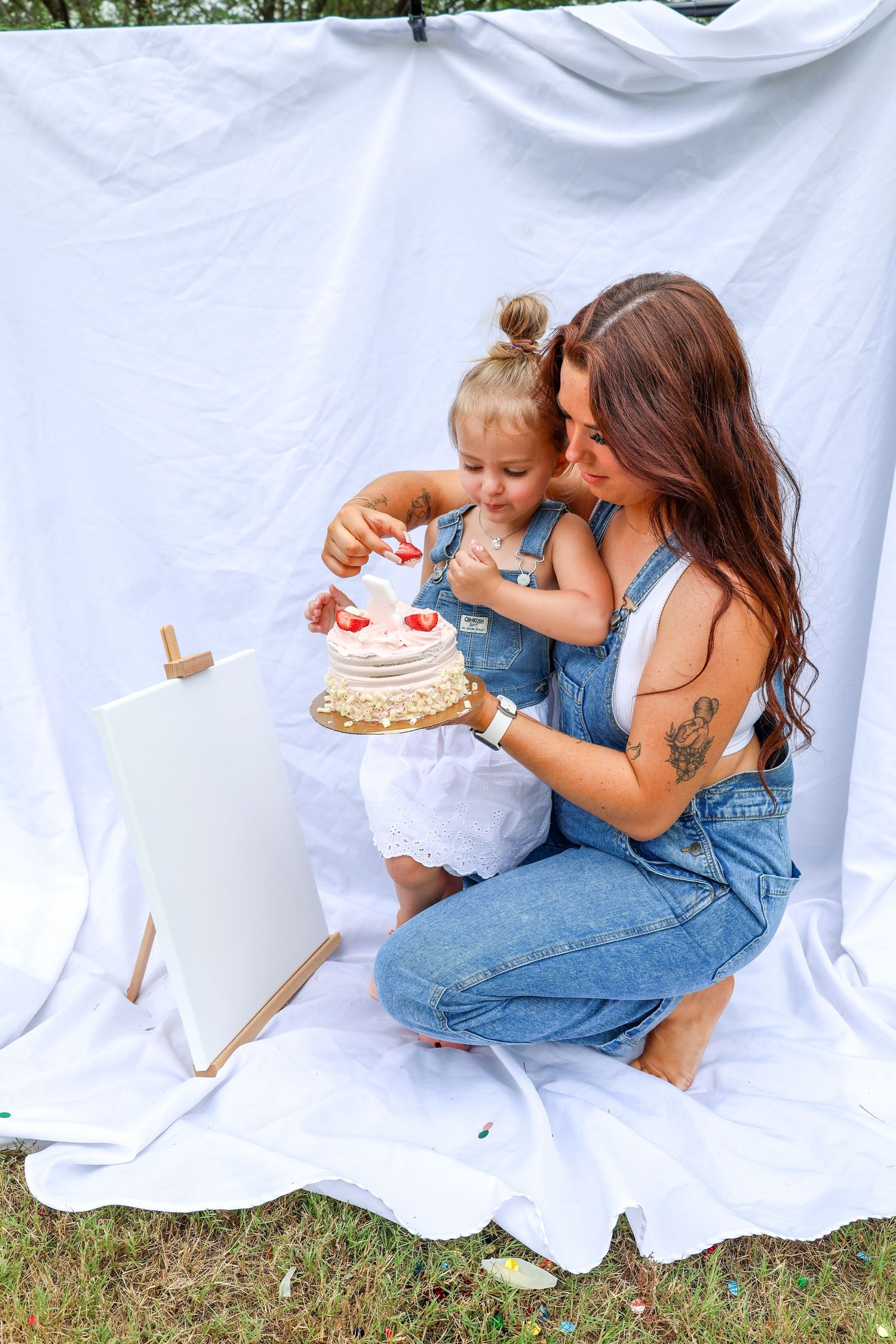 White draped cloth backdrop; child in a white dress and woman with red brunette hair wearing a white top and overalls while holding a cake