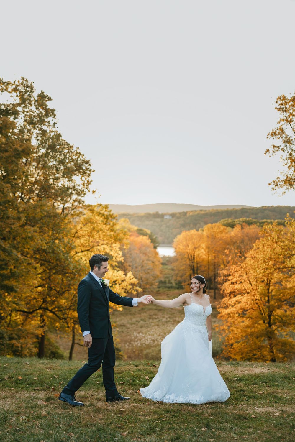 A couple in wedding attire walks through a golden autumn field at sunset with trees and rolling hills in the background.
