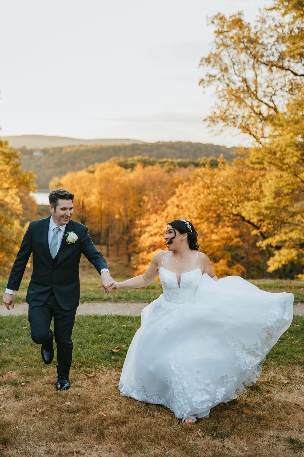 Bride and groom playfully run through a golden autumn field at sunset with fall foliage in the background.